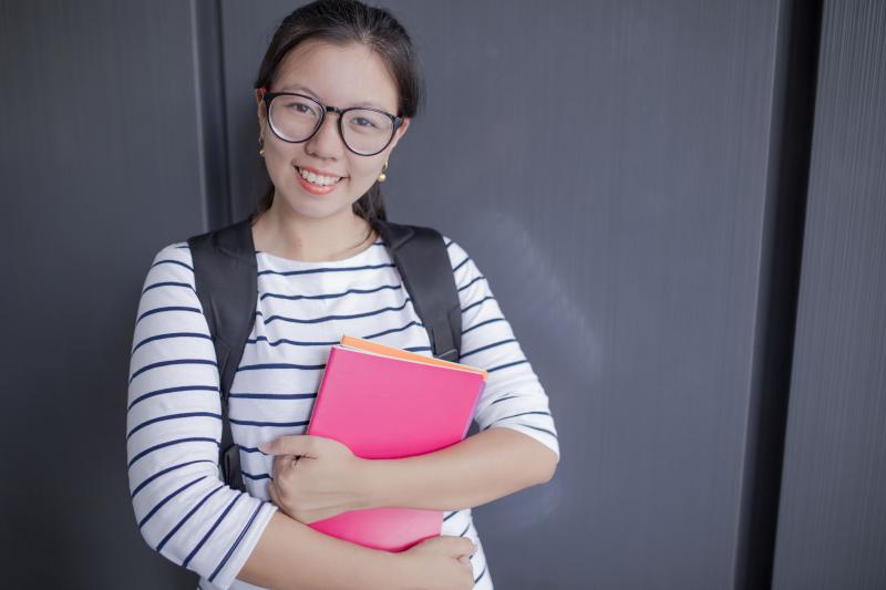 An East Asian girl smiling while wearing glasses, a striped shirt, a backpack and holding a pink book.