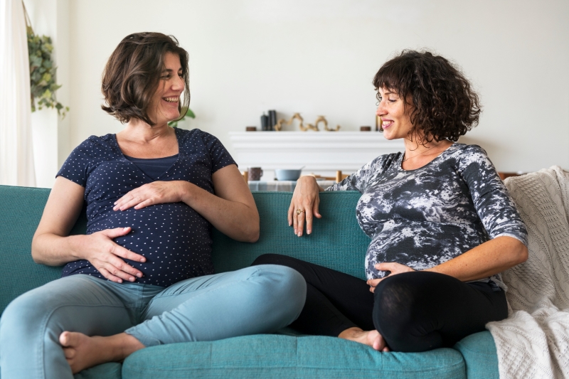 Two pregnant woman talking on a blue couch.