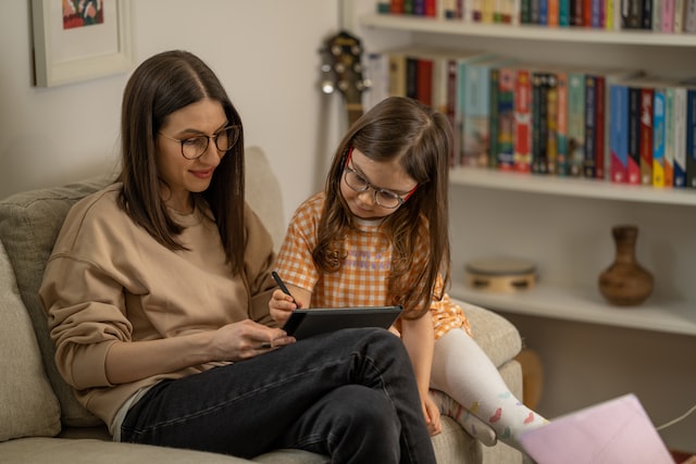 A woman and a school-age child sitting on a couch and looking at an iPad. They are both wearing glasses.