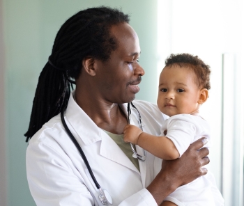 A Black doctor holding a baby. He has his hair tied back and is wearing a lab coat.
