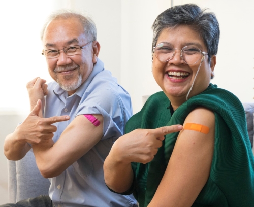 An elderly man and woman sitting on a couch while pointing to the band aid on their arm and smiling.