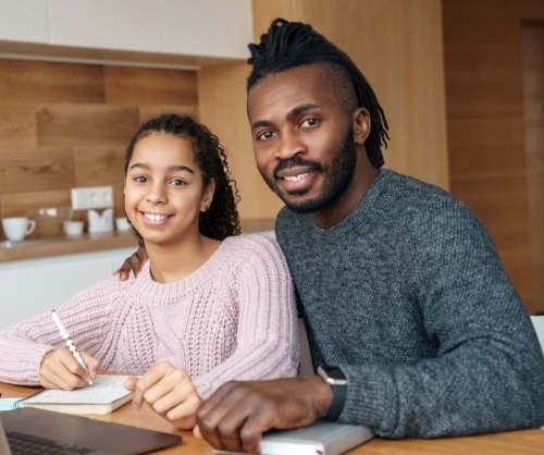 A Black man sitting at a table in a kitchen with a teenage girl working on a laptop.