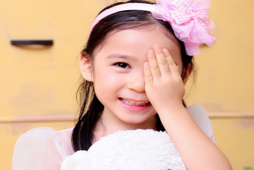 An East Asian girl smiling while holding her hand over her left eye. She is holding a white teddy bear.