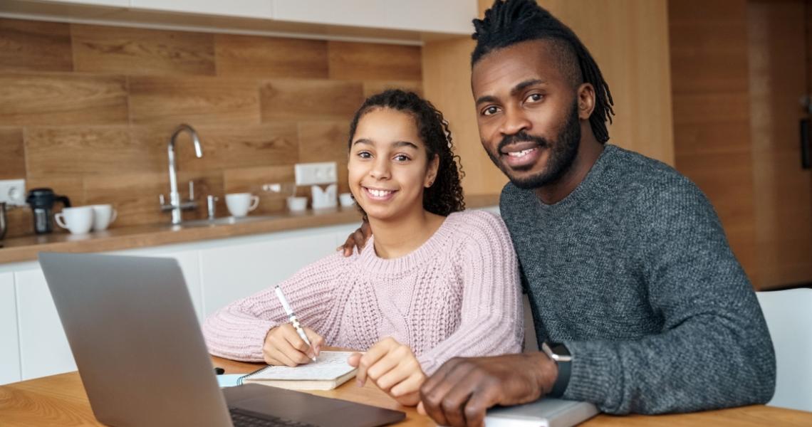A Black man sitting at a table in a kitchen with a teenage girl working on a laptop.