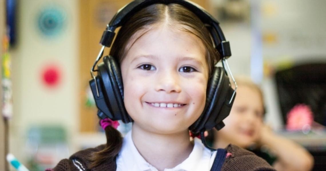 A school-aged girl in a classroom smiling. She is holding a pencil and wearing black headphones.