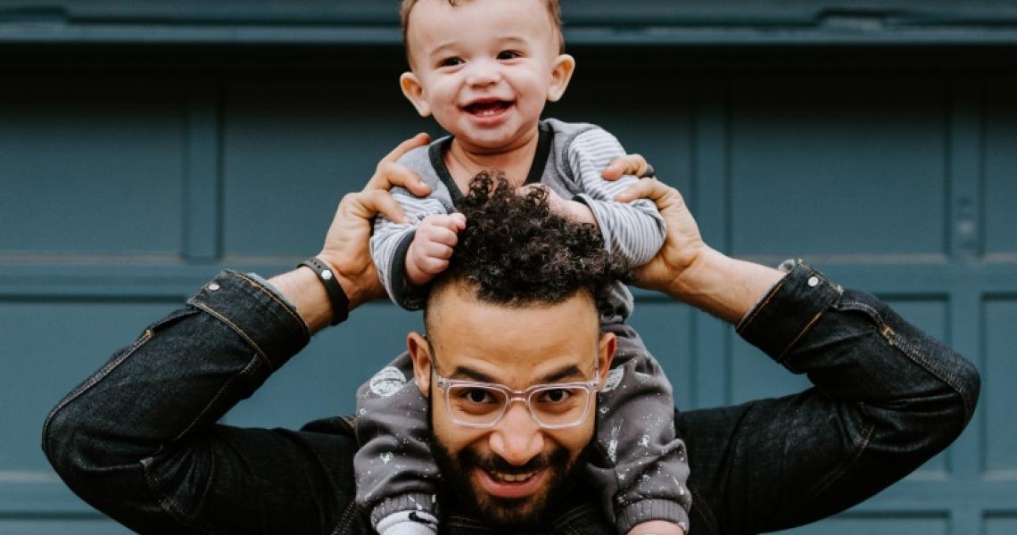 A Brown man smiling while holding a smiling baby on his shoulders infront of a blue garage door.