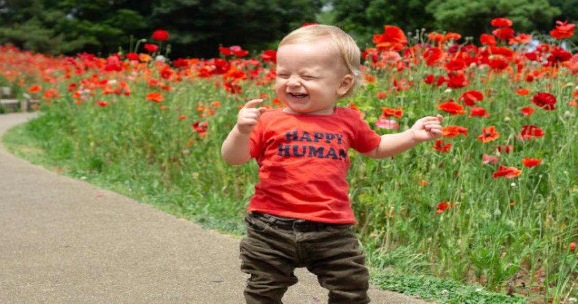 A young infant boy standing infront of flowers and smiling. He is wearing a bright orange shirt with &quot;Happy Human&quot; written on it.