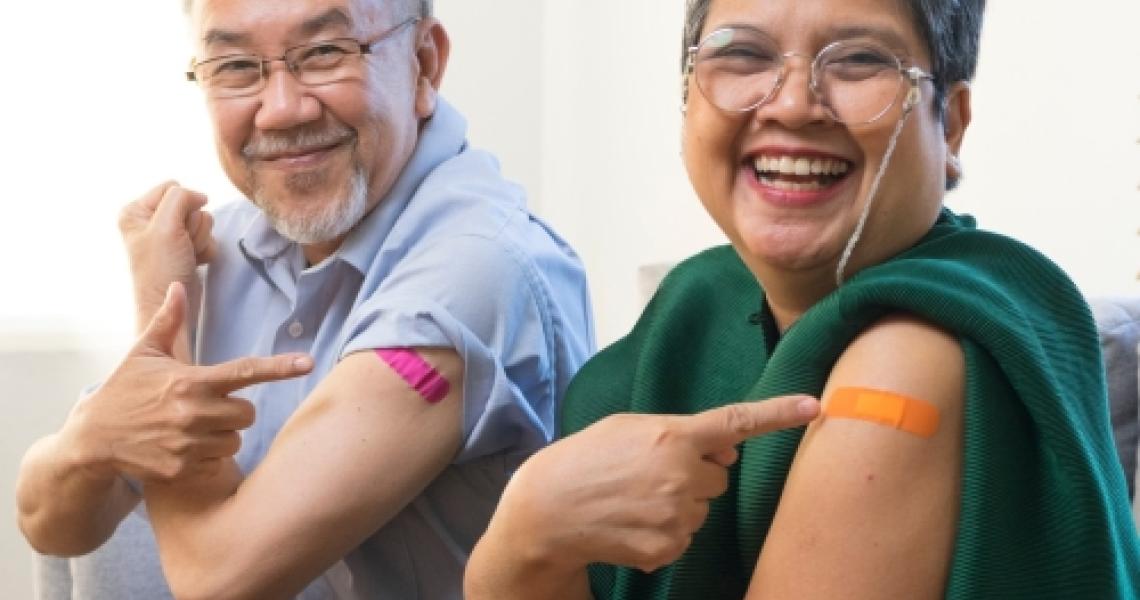 An elderly man and woman sitting on a couch while pointing to the band aid on their arm and smiling.