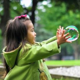 An East Asian girl with a green coat, playing with bubbles in a park.