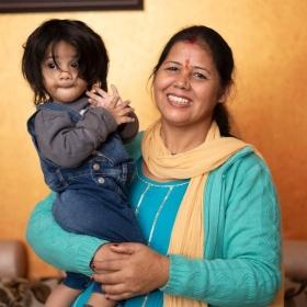 A South Asian woman smiling while holding her infant child. They are standing infront of an orange wall.