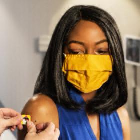 A Brown woman wearing a yellow mask is receiving a band aid after getting a vaccine.