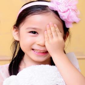An East Asian girl smiling while holding her hand over her left eye. She is holding a white teddy bear.