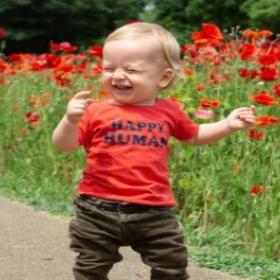 A young infant boy standing infront of flowers and smiling. He is wearing a bright orange shirt with "Happy Human" written on it.