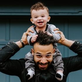 A Brown man smiling while holding a smiling baby on his shoulders infront of a blue garage door.