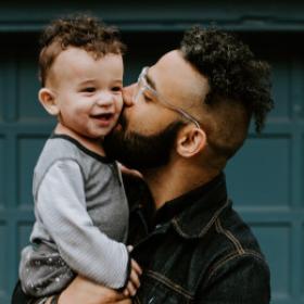 A Brown man holding a smiling baby in his arms and kissing his cheeck infront of a blue garage door.