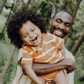 A Black man smiling while holding a Black toddler laughing.